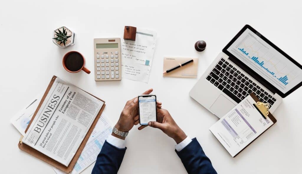 a flat lay of a newspaper, laptop, calculator, and hands holding a cell phone